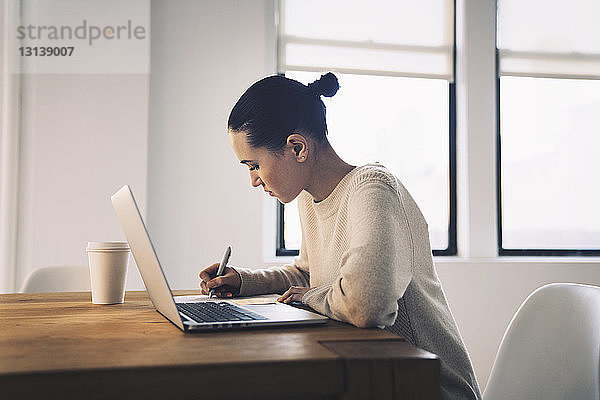 Geschäftsfrau schreibt  während sie mit einem Laptop am Schreibtisch im Büro sitzt
