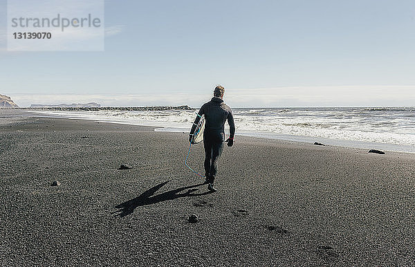 Rückansicht eines Mannes mit Surfbrett  der am schwarzen Strand am sonnigen Tag gegen den Himmel läuft