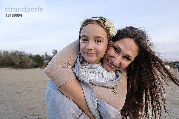Porträt einer lächelnden Mutter  die ihre Tochter umarmt  während sie am Strand gegen den Himmel steht