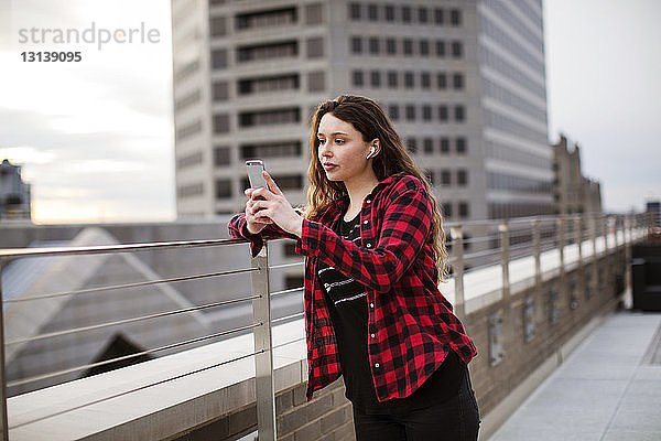 Junge Frau benutzt ein Smartphone  während sie auf der Terrasse am Geländer steht