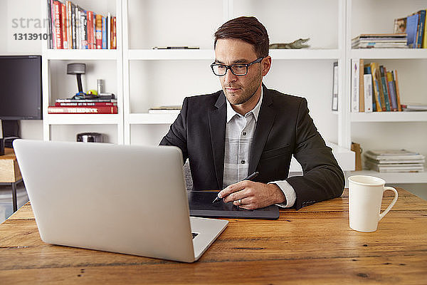 Geschäftsmann mit Grafiktablett und Laptop-Computer am Holztisch im Heimbüro