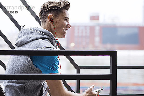 Seitenansicht eines glücklichen männlichen Sportlers  der über ein Smartphone auf der Treppe Musik hört