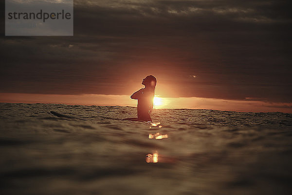 Silhouette einer Frau  die bei Sonnenuntergang auf dem Meer vor bewölktem Himmel surft