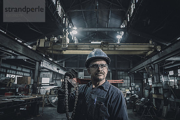 Männlicher Arbeiter  der in der Fabrik stehend ein Arbeitsgerät trägt und dabei wegschaut