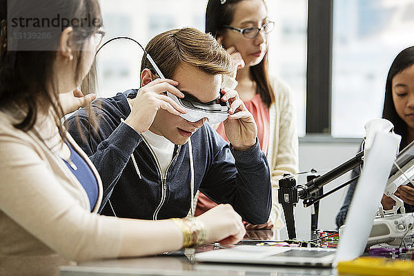 Männlicher Student trägt eine Virtual-Reality-Brille  während er im Klassenzimmer bei Freunden sitzt