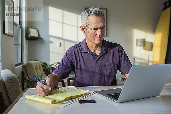 Reifer Mann schreibt auf Notizblock  während er zu Hause einen Laptop-Computer benutzt