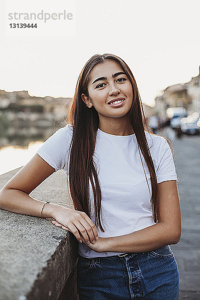 Porträt einer lächelnden jungen Frau  die an einer Stützmauer in der Stadt steht