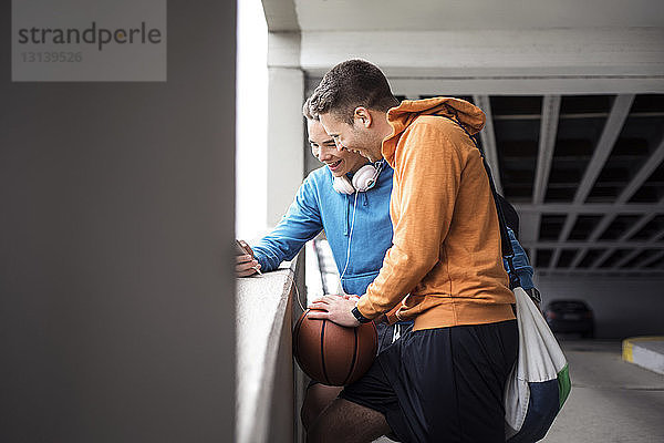 Fröhliche männliche Freunde mit Basketball mit Smartphone auf dem Parkplatz