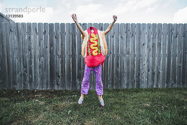 Mädchen mit erhobenen Armen springt im Hot-Dog-Kostüm gegen den Zaun im Hof