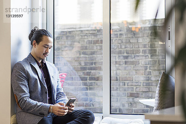 Geschäftsmann benutzt Mobiltelefon  während er im Büro auf einem Fensterplatz in einer Nische sitzt