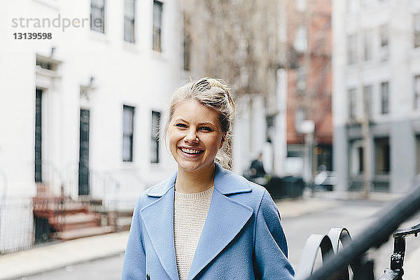 Porträt einer fröhlichen jungen Frau in Trenchcoat  die vor Gebäuden in der Stadt steht