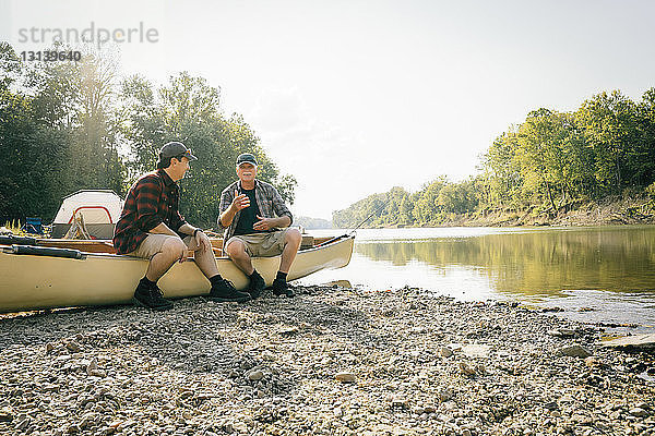 Männliche Freunde unterhalten sich  während sie auf einem Boot gegen den Himmel sitzen