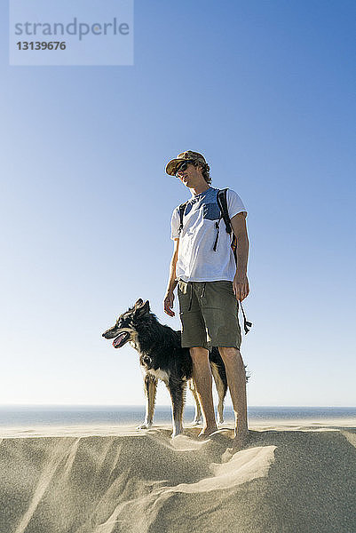 Mann mit Hund steht am Strand vor klarem blauen Himmel