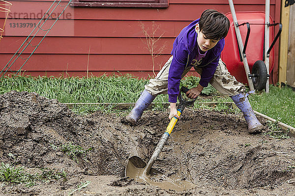 Junge in voller Länge gräbt Feld mit Spaten während der Gartenarbeit im Hinterhof