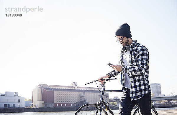 Mann benutzt Telefon  während er sein Fahrrad am Kanal gegen den klaren Himmel hält