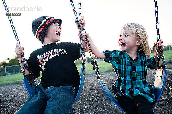 Fröhliche Geschwister genießen auf dem Spielplatz die Schaukel gegen den Himmel