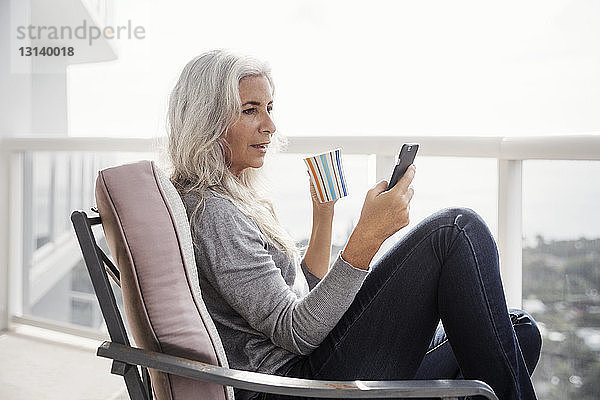 Seitenansicht einer reifen Frau  die ein Smartphone benutzt  während sie auf dem Balkon Kaffee trinkt