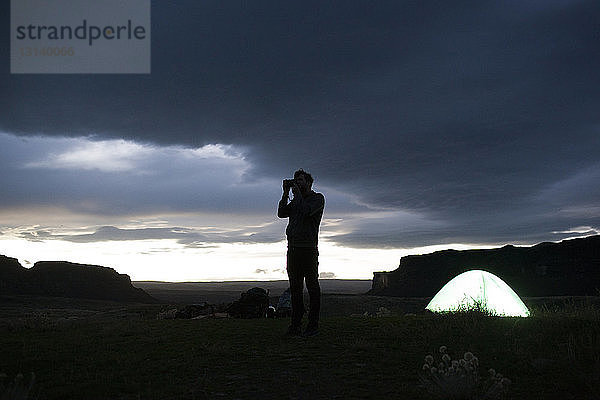 Scherenschnitt-Wanderer fotografiert  während er in der Dämmerung auf dem Feld im Zelt vor stürmischen Wolken steht