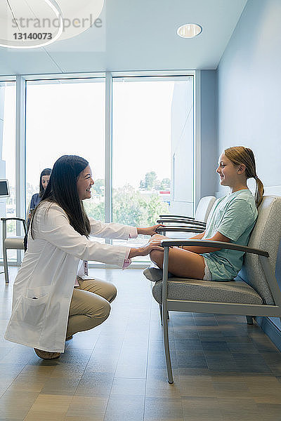 Fröhlicher Kinderarzt spricht mit Mädchen  während er im Wartezimmer des Krankenhauses hockt