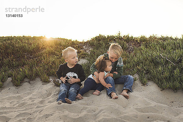 Geschwister in voller Länge  die bei Sonnenuntergang im Sand am Strand vor klarem Himmel sitzen