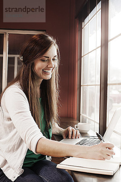 Glückliche Frau schreibt in Buch  während sie im Café einen Laptop benutzt