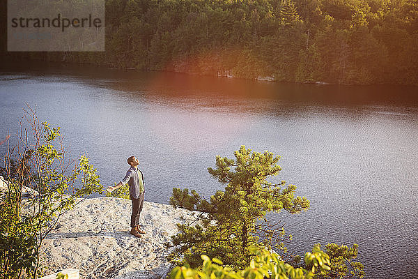 Hochwinkelaufnahme eines jungen Mannes  der mit ausgestreckten Armen auf einem Felsen mit Blick auf den See steht