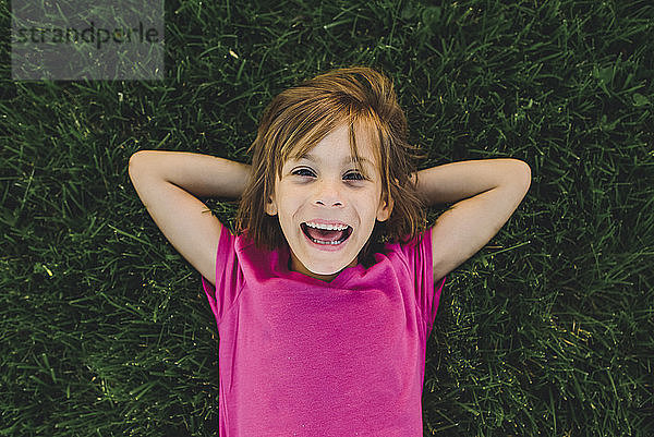 Überkopf-Porträt eines fröhlichen Mädchens mit Händen hinter dem Kopf  das auf einem Grasfeld liegt