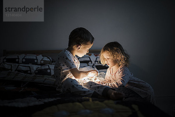 Geschwister mit beleuchteten Lichterketten  die zu Hause auf dem Bett sitzen