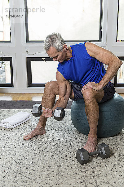 Ein muskulöser  reifer Mann in voller Länge  der eine Hantel hebt  während er zu Hause auf einem Fitnessball sitzt