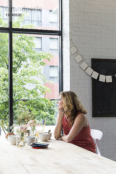 Nachdenkliche Frau mit Hand am Kinn  die durch das Fenster schaut  während sie im Café sitzt