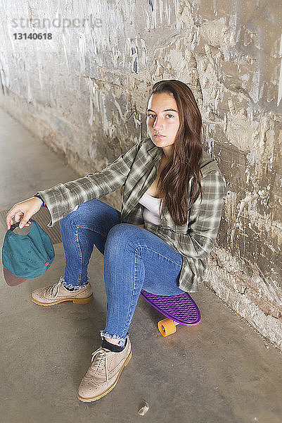 Porträt eines selbstbewussten Schülers  der auf einem Skateboard vor einer verwitterten Wand sitzt