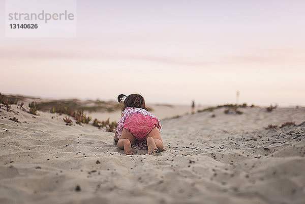 Rückansicht eines kleinen Mädchens  das bei Sonnenuntergang auf Sand am Strand gegen den Himmel krabbelt