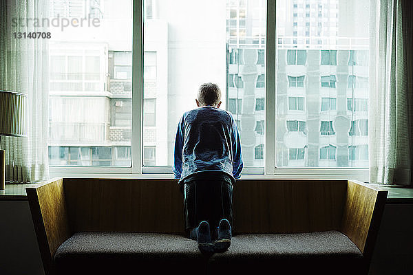 Rückansicht eines auf dem Sofa knienden Jungen  der zu Hause durch ein Fenster blickt