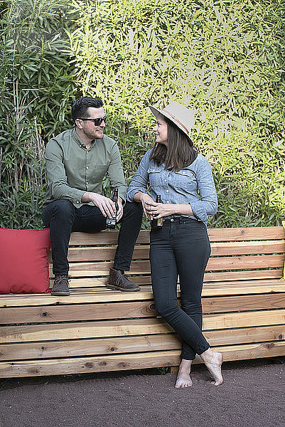 Paar hält Bierflaschen in der Hand und unterhält sich auf einer Holzbank im Hinterhof