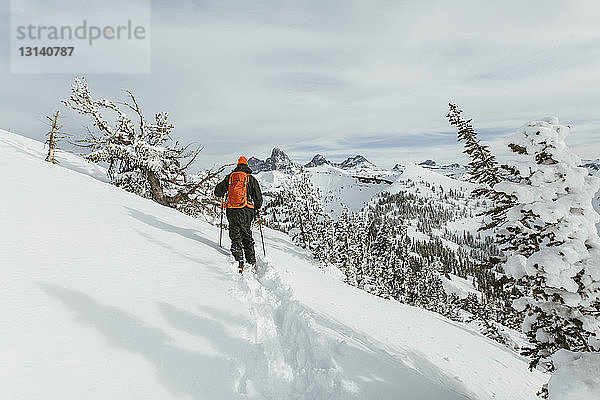 Rückansicht eines Wanderers mit Rucksack und Skistöcken beim Gehen auf einem schneebedeckten Berg