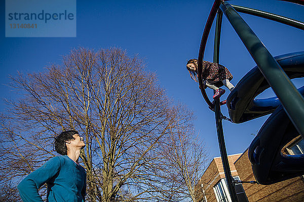 Niedrigwinkelansicht eines Vaters  der seine Tochter beim Klettern auf Außenspielgeräten auf dem Spielplatz beobachtet