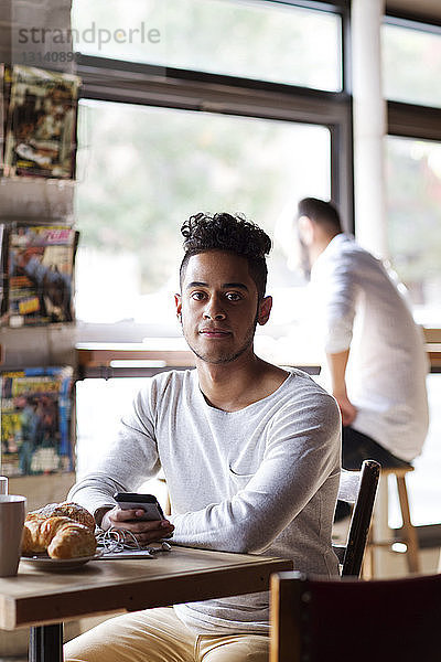 Porträt eines jungen Mannes mit Mobiltelefon und Croissant im Cafe sitzend