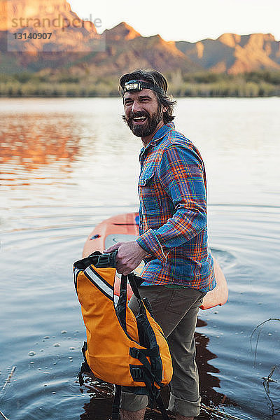 Seitenansicht Porträt eines glücklichen Mannes mit Paddelbrett am Seeufer stehend