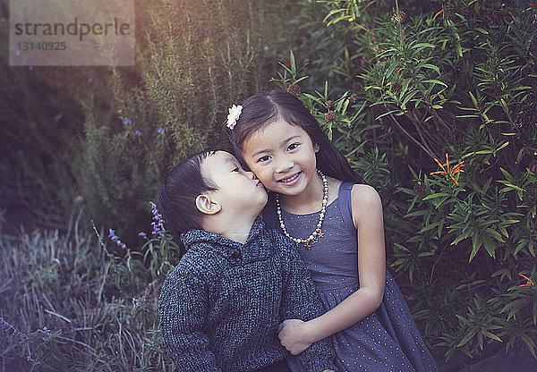 Porträt einer niedlichen Schwester mit einem Bruder  der sie im Park gegen Pflanzen küsst