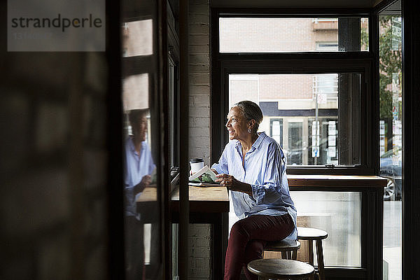 Lächelnde Frau liest Zeitung  während sie durch das Fenster am Tisch im Cafe schaut