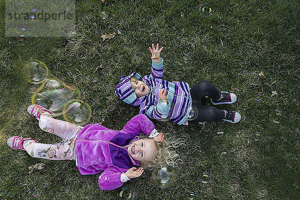 Draufsicht auf Geschwister  die mit Blasen spielen  während sie auf einem Grasfeld liegen