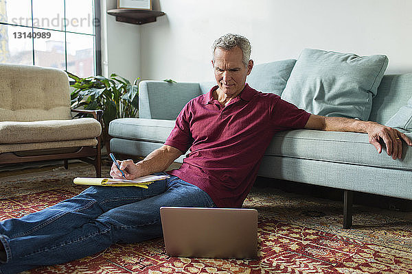 Ernsthaft reifer Mann  der einen Laptop benutzt  während er zu Hause im Wohnzimmer neben dem Sofa auf dem Teppich sitzt