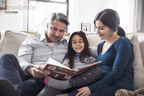 Mädchen mit Vater und Mutter liest Buch  während sie zu Hause auf dem Sofa sitzen