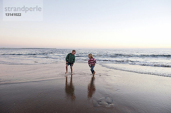 Verspielte Geschwister  die bei Sonnenuntergang am Strand gegen den Himmel laufen