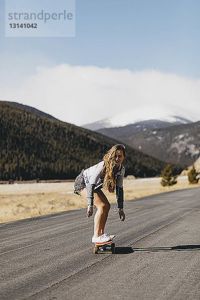 Unbeschwerte junge Frau fährt bei Sonnenschein in voller Länge Skateboard auf der Straße