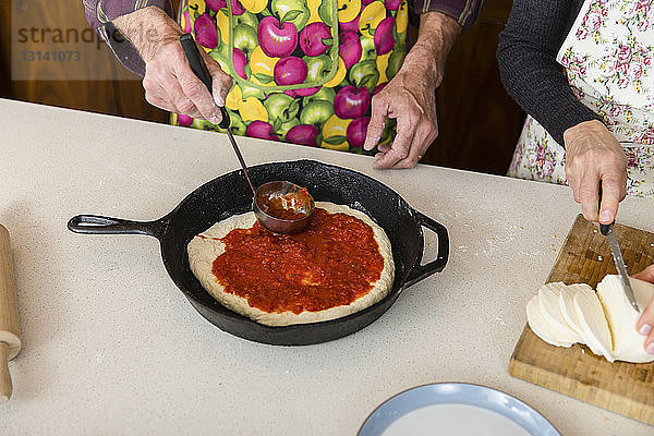 Mittelteil eines Mannes  der in der Bratpfanne Zutaten auf den Pizzateig aufträgt  während die Frau zu Hause Käse schneidet