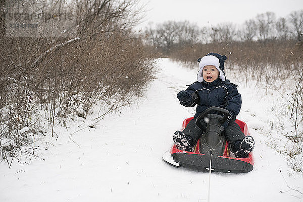 Ein süßer kleiner Junge sitzt in voller Länge auf einem Schlitten mitten im Schnee