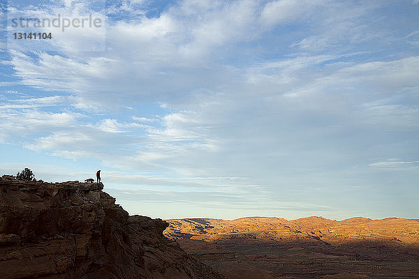 Fernansicht eines Silhouettenwanderers mit Hund  der auf dem Gipfel eines Berges vor bewölktem Himmel steht