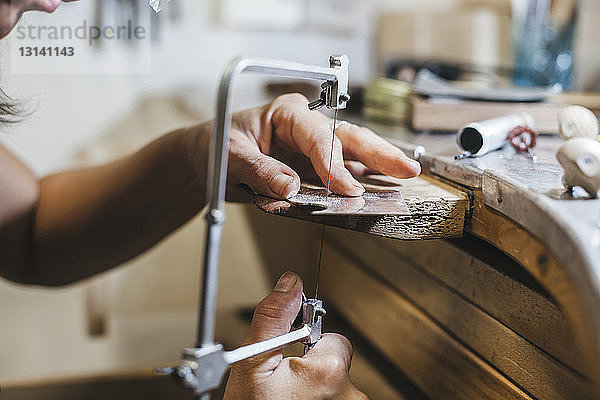 Geschnittene Hände einer Kunsthandwerkerin mit einer Handsäge bei der Herstellung von Schmuck auf einem Holztisch in einer Werkstatt