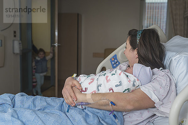 Mutter hält neugeborenen Sohn im Arm und sieht Tochter beim Eintritt in die Krankenhausstation an  während sie sich auf dem Bett ausruht